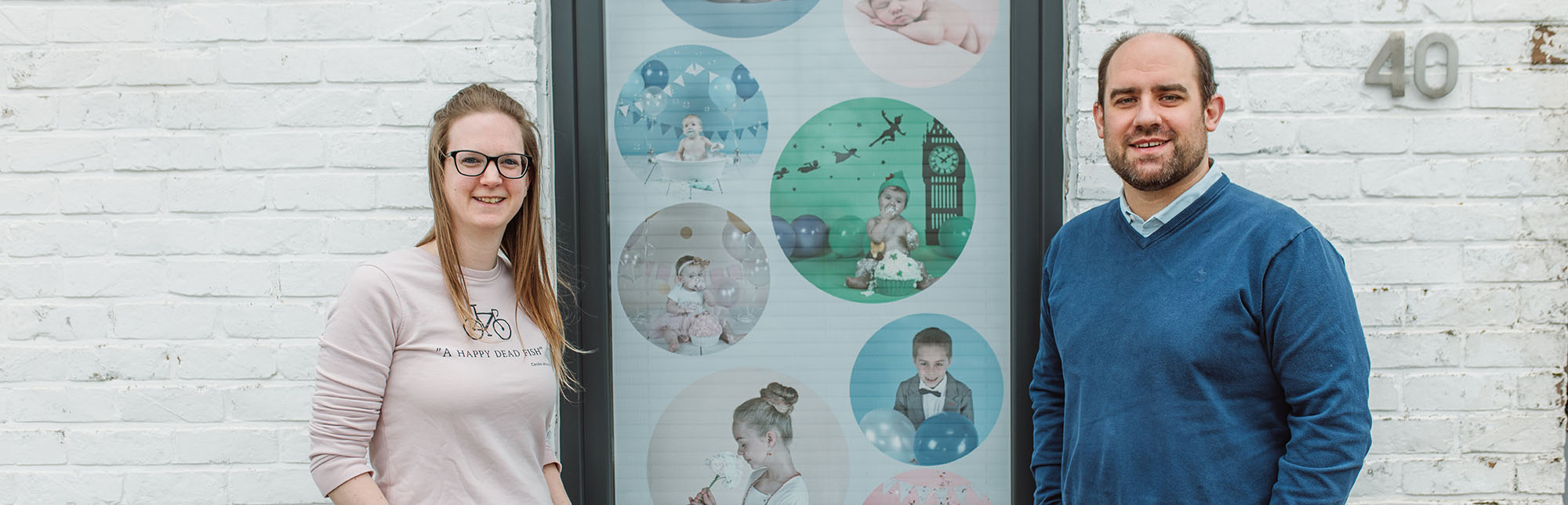 Portret van Pieter en Tine die voor hun gevel staan aan een banner met kinderfoto's, zaakvoerders van Studio Gaston.