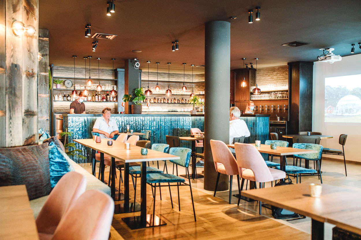 Prachtig interieur met sfeervolle verlichting boen de toog. De toog is afgewerkt met een blauwe tegel. Ervoor staan tafels met roze stoelen.