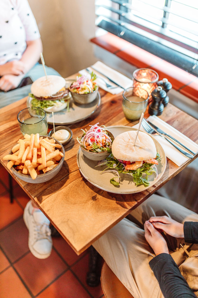 Close up van een tafel met 2 hamburgers met slaatje, en een kom met frietjes.