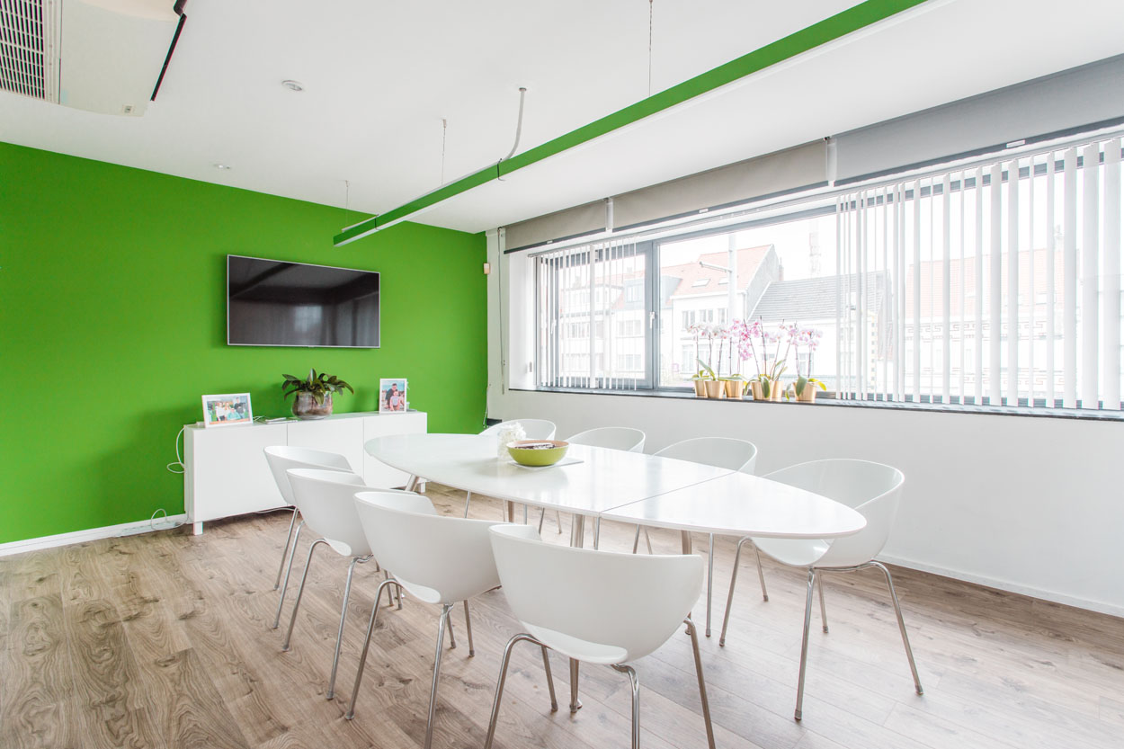 Een vergaderzaal met witte grote tafel met 8 witte kuipstoelen aan en achteraan een groene muur.