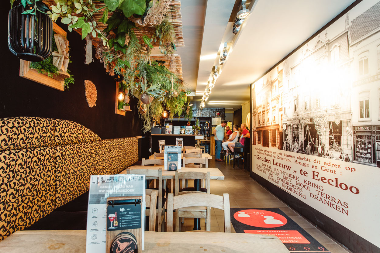 Het interieur. Donkere muur met bovenaan kunstplanten aan het plafond. Houten stoelen en tafels.