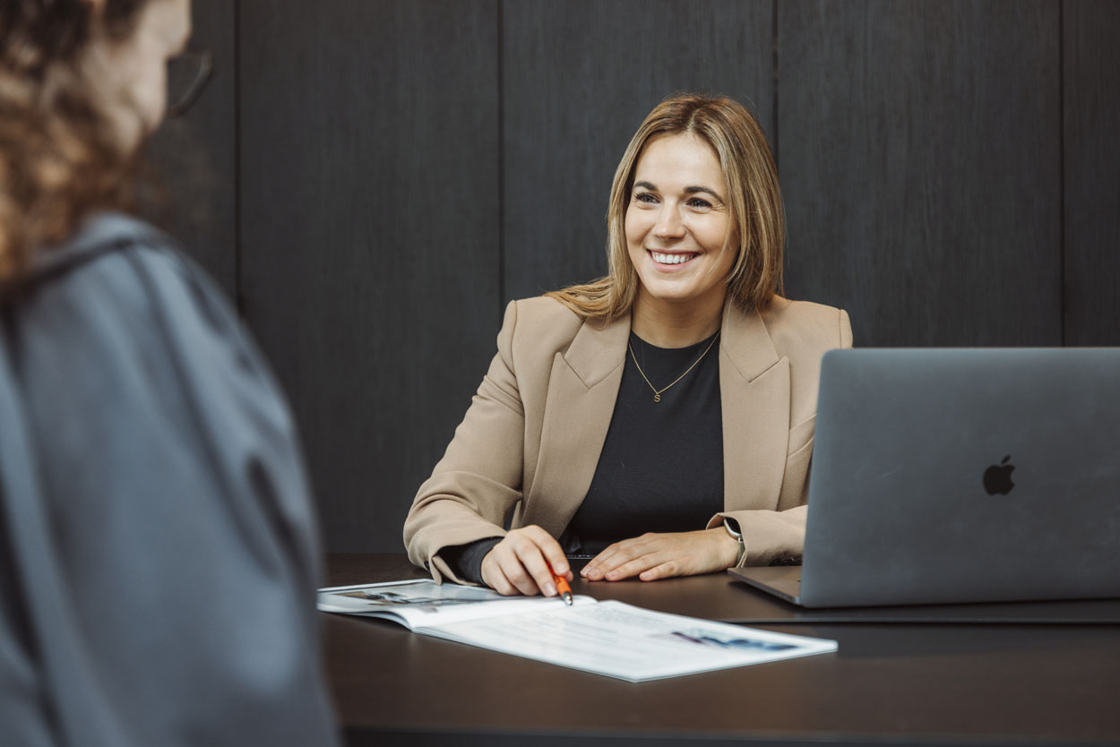 Stefanie wijst met haar stylo naar een document op tafel en glimlacht naar de klant.