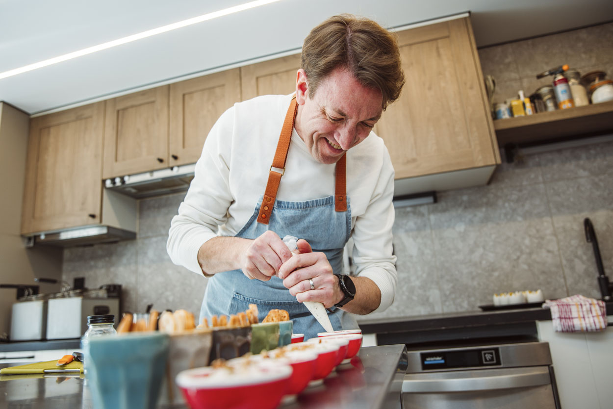 Dirk is het eten aan het klaarmaken. Hij staat glimlachend potjes te vullen met een spuitzak.